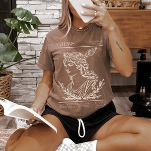 May include: A brown t-shirt with a graphic of Artemis, the Greek goddess of the hunt. The graphic includes a line drawing of Artemis with a bow and arrow, surrounded by laurel leaves. The text "ARTEMIS", "GODDESS OF THE HUNT", "LADY OF THE WILD THINGS, PROTECTOR OF THE YOUNG", and "Daughter of Zeus & Leto" is printed on the shirt.
