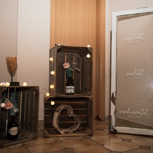 May include: Three wooden crates with string lights, a heart-shaped wooden cutout, and a bottle of liquor. A white door with the words "verliebt", "verlobt", and "verheiratet" with dates written on it.
