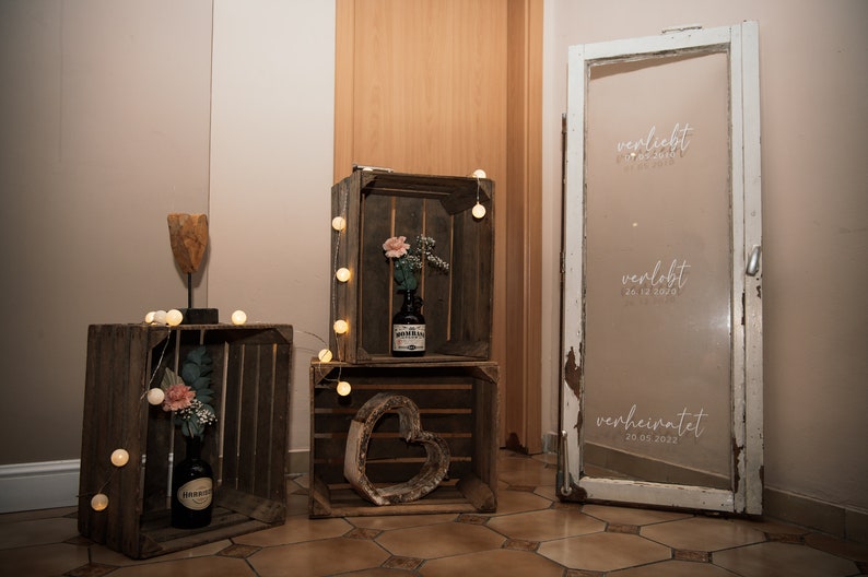 May include: Three wooden crates with string lights, a heart-shaped wooden cutout, and a bottle of liquor. A white door with the words "verliebt", "verlobt", and "verheiratet" with dates written on it.