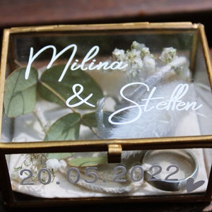 May include: A glass box with a gold frame, containing a white feather, greenery, and two rings. The box is engraved with the text "Milina & Steffen" and the date "20.05.2022".