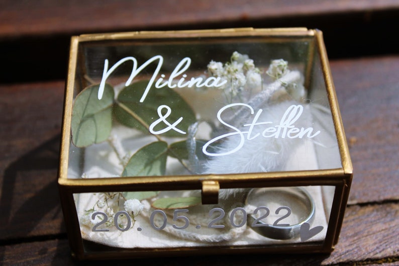 May include: A glass box with a gold frame, containing a white feather, greenery, and two rings. The box is engraved with the text "Milina & Steffen" and the date "20.05.2022".