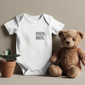 Può includere: Un body bianco per neonato con maniche corte e il testo "FRECHS DACHs." stampato sul davanti. Un orsacchiotto di peluche marrone è seduto accanto al body.