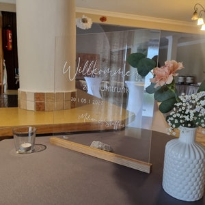 May include: A clear acrylic sign with the text "Willkommen zum Umtrunk 20 | 05 | 2022 Melina & Steffen" written in white. The sign is on a wooden stand and is decorated with white and pink flowers.