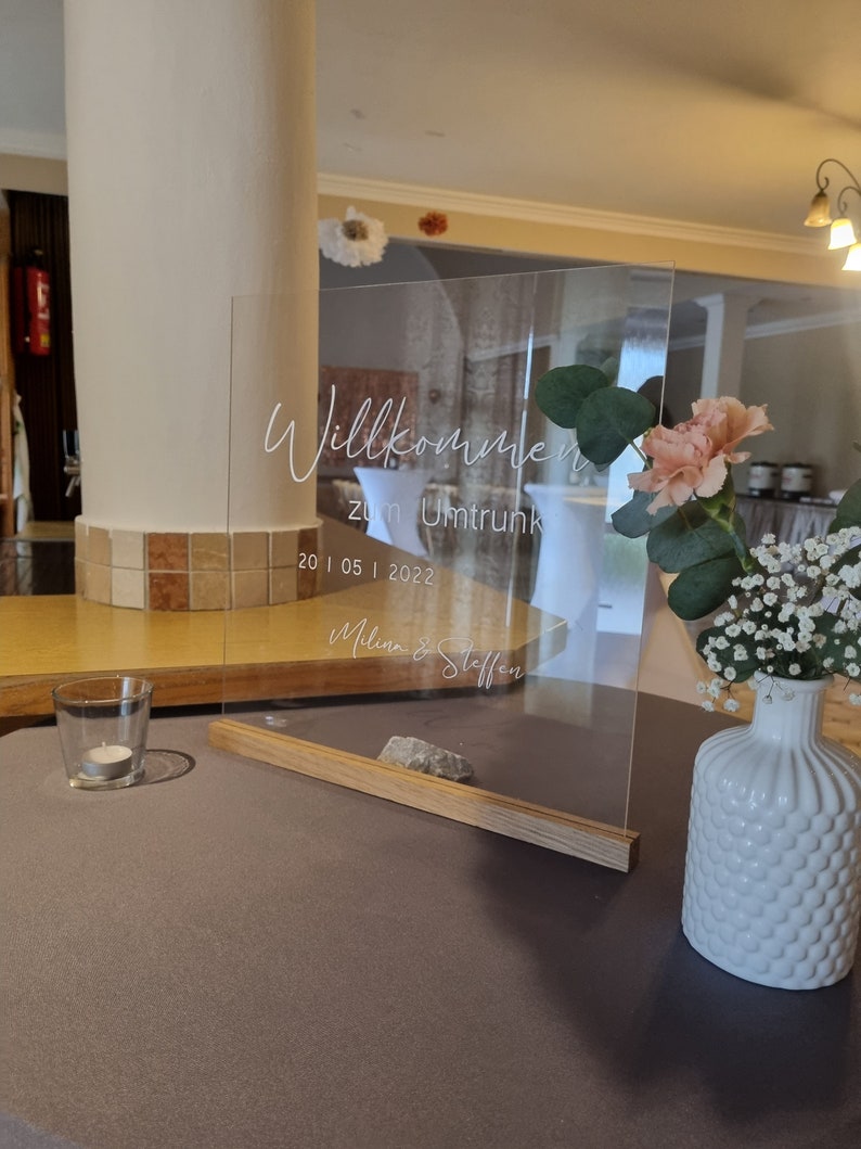 May include: A clear acrylic sign with the text "Willkommen zum Umtrunk 20 | 05 | 2022 Melina & Steffen" written in white. The sign is on a wooden stand and is decorated with white and pink flowers.