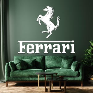 Può includere: Logo Ferrari bianco con un cavallino rampante sopra il nome del marchio in un font moderno. Il logo è montato su una parete verde scuro sopra un divano in velluto verde con gambe in legno. Un tavolino e una pianta in vaso sono di fronte al divano.