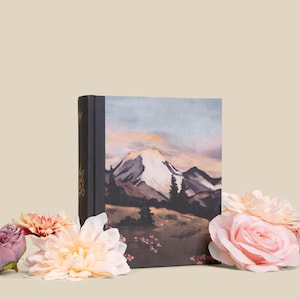 May include: A hardcover book with a mountain landscape painting on the cover, featuring a snow-capped peak under a pastel sky. The book is surrounded by pink and peach-colored flowers, creating a soft, natural aesthetic.