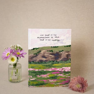 May include: A hardcover book featuring a painted landscape of a mountain and a field of pink flowers. The text "The God of the mountain is the God of the valley" is written in black script on the sky portion of the cover art.