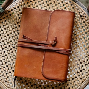 May include: A brown leather journal with a wrap-around tie closure. The journal is rectangular and has a smooth, textured surface. The tie is made of the same leather and wraps around the journal to secure it.