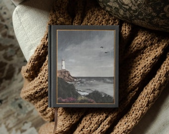 May include: A hardcover book with a coastal landscape painting on the cover, featuring a lighthouse on a cliffside. The book has a dark gray cover with a gold border and a brown ribbon bookmark. It rests on a tan knitted blanket.