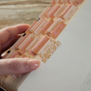 May include: Pink and gold book tabs with floral designs and text. The tabs are attached to the pages of a notebook or journal. The word "ACTS" is visible on one tab.