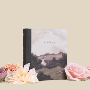May include: A hardback book with a landscape painting of a church and hills. The spine is black, and the cover features the text "The Word of God". Pink and peach flowers are arranged around the book.