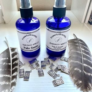 Può includere: Due flaconi spray in vetro blu con etichette bianche. Le etichette riportano "Powerful Bracelet" e "Smudge Spray Palo Santo" e "Money Mist". I flaconi sono appoggiati su una superficie bianca con ciondoli argentati sparsi intorno.