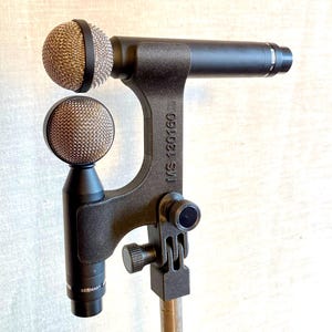 May include: A black stereo microphone setup with two mesh-covered microphone heads. The stand is black with the text "MS 130160" and "GERMANY" printed on the microphone body. The microphone is designed for professional audio recording.