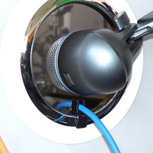 Puede incluir: Un micrófono negro con un cable azul conectado está colocado dentro del agujero de sonido de un tambor. El micrófono está montado en un soporte y está inclinado hacia el interior del tambor.
