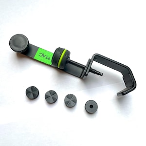 Puede incluir: Abrazadera de soporte de micrófono negro con una perilla verde y negra. La abrazadera tiene un brazo de metal negro con un tornillo y una perilla de plástico negro. La abrazadera está unida a una base de metal negro. Hay cuatro patas de plástico negro y una perilla de plástico negro en la base. La abrazadera está etiquetada con "MAC" en marcador verde.