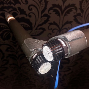 Peut inclure: Gros plan d'un microphone à condensateur à double élément noir avec un pare-brise en mousse. Le microphone est monté sur un support noir avec un câble bleu.
