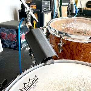 Op de afbeelding: Een close-up van een drumstel met een microfoon bevestigd aan de trommel. De trommel heeft een witte kop met het "Remo"-logo. De microfoon is zwart en verbonden met een blauwe kabel. De trommel is bruin met een houtnerf afwerking.