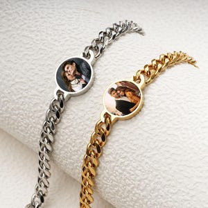 Può includere: Due braccialetti con catene a maglia. Uno è color argento, l'altro dorato. Ognuno presenta un ciondolo circolare con una fotografia a colori di una coppia. I braccialetti sono esposti su una superficie bianca strutturata.