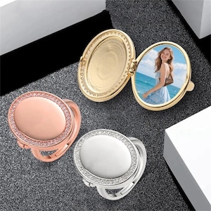 May include: Three oval-shaped locket rings in gold, rose gold, and silver. The gold locket is open, displaying a photo of a person. The rings have a decorative border of small, clear stones. The rose gold and silver rings are closed.