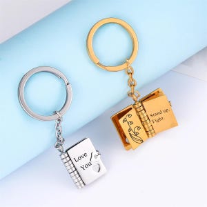 May include: Two keychains, one silver and one gold, each featuring a miniature book pendant. The silver keychain reads "Love You" with a heart cutout. The gold keychain has the words "Stand up. Fight." and a floral design.