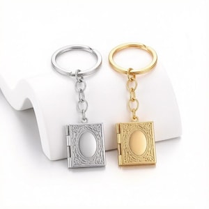 May include: Two metal keychains featuring a small, square locket pendant with an ornate, embossed floral border and a smooth, oval center. One keychain is finished in a bright silver tone, while the other is finished in a polished gold tone.
