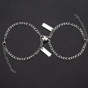 Puede incluir: Dos pulseras de cadena plateadas con dijes en forma de coraz&oacute;n y etiquetas rectangulares. Las pulseras est&aacute;n unidas por los dijes en forma de coraz&oacute;n.