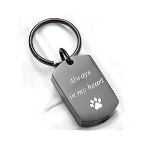 Puede incluir: Llavero con placa de identificación para perro de color gris oscuro con un anillo negro. La placa tiene las palabras "Always in my heart" y una huella de pata blanca. Un artículo conmemorativo.