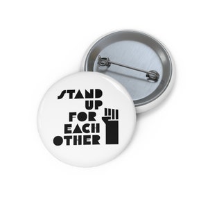 May include: White button with black text that reads "Stand Up For Each Other" and a black fist raised in the air.