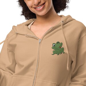 May include: A tan zip-up hoodie with a green frog embroidered on the left chest. The frog is winking and sticking out its tongue.