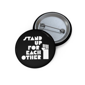 May include: Black button with white text that reads "Stand Up For Each Other" and a raised fist graphic.