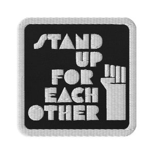May include: Black and white embroidered patch with the text "Stand Up For Each Other" and a raised fist.