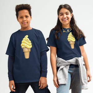 May include: Two children in navy blue t-shirts featuring a yellow ice cream cone design. One child has a long-sleeved top underneath. Both wear jeans and trainers. The background is a blend of white and brown.