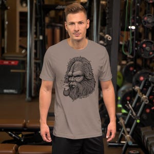 May include: A gray t-shirt with a black and white illustration of a creature with a beard smoking a pipe. The creature has a large head and a long beard. The text "Thought you were alone?" is printed below the illustration.