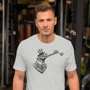 May include: Light gray t-shirt featuring a black and white illustration of a crocodile gentleman wearing a top hat and suit, smoking a pipe. The graphic is centered on the chest. The shirt is short-sleeved and has a crew neck.