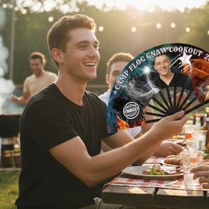 May include: A black hand fan with a photo of a man and the text "CAMP FLOG GNAW COOKOUT 2025" held by a person at an outdoor cookout. The fan has a black handle and a colorful design with a star.