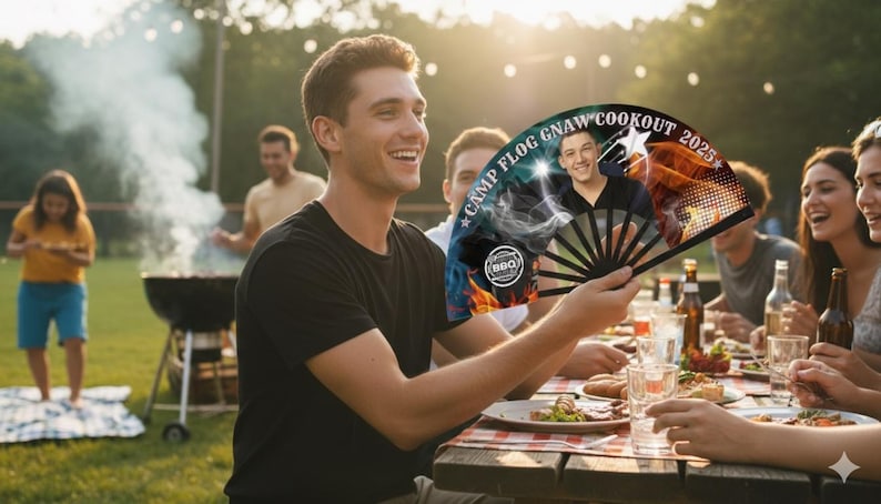May include: A black hand fan with a photo of a man and the text "CAMP FLOG GNAW COOKOUT 2025" held by a person at an outdoor cookout. The fan has a black handle and a colorful design with a star.