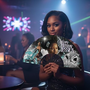 May include: A woman holding a decorative fan with the text "Happy Birthday Jessica" in a nightclub setting. The fan features a photo and playing card graphics. The background includes other people, lights, and tables.