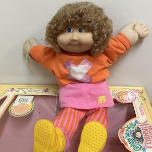以下が含まれることがあります： カールした茶色の髪と青い目のキャベツパッチキッズ人形。人形は、白いハートが付いたオレンジ色のセーター、ピンクのスカート、オレンジとピンクのストライプのパンツを着ています。 黄色の靴。