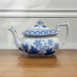 May include: White ceramic teapot with a blue floral design. The teapot features a curved spout, a curved handle, and a lid with a decorative finial. The body of the teapot is decorated with blue flowers and leaves.