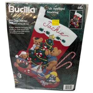 Könnte beinhalten: Ein Bucilla Teddy Bear Christmas Filz-Applikations-Strumpf-Set in einer Kunststoffverpackung. Der rote Strumpf hat eine weiße Manschette mit dem Namen Jackie, verziert mit Teddybären, einem Weihnachtsbaum, Spielzeug und Zuckerstangen, 38,1 cm.