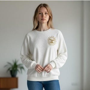 Peut inclure: Un sweat-shirt blanc à manches longues et col rond, confectionné dans un tissu texturé semblable à de la polaire. Le vêtement présente un petit motif doré représentant un soleil avec un visage, imprimé sur la poitrine gauche. Bords-côtes.