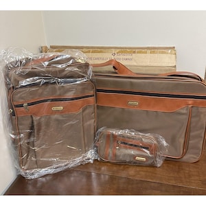 Pode incluir: Um conjunto de bagagem Samsonite vintage em castanho e bege. O conjunto inclui uma mala grande, uma pasta menor e uma bolsa de cosméticos. Cada peça tem detalhes em couro castanho e zíperes dourados. A bagagem está embalada em plástico.