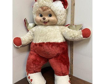 May include: A plush doll with a cream-colored fuzzy hood and body, red plush pants, and red-tipped hands. The doll features gold wings, white boots with red heart accents, and a plastic face. A wooden ruler shows the doll is 26 inches tall.