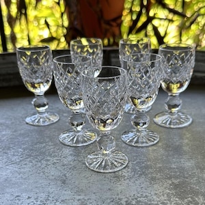 May include: A set of eight clear crystal wine glasses. Each glass features a diamond-cut pattern on the bowl, a round stem, and a starburst base. The glasses are arranged on a gray surface with a blurred green background.