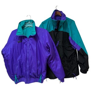 May include: Two vintage Columbia jackets. One is purple with teal accents, the other is black, teal, and purple. Both have zip closures, pockets, and drawstrings. The jackets are hanging on hangers against a white background.