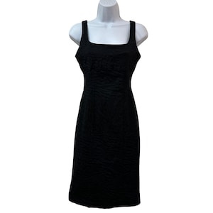 May include: A black, textured sheath dress with a square neckline and thick straps. The dress is displayed on a white mannequin. The fabric has a subtle, all-over pattern, and the dress falls to the knee.
