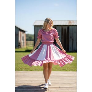 May include: A two-piece outfit consisting of a dusty rose short-sleeved top with a ruffled neckline and a matching skirt. The skirt features alternating vertical panels of solid white and a dusty rose fabric with a subtle white polka dot pattern.