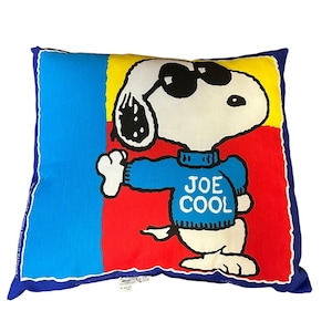 Puede incluir: Cojín cuadrado con Snoopy con gafas de sol y un suéter azul con la inscripción "JOE COOL". El cojín tiene un borde azul y un fondo colorido de rojo, amarillo y azul.