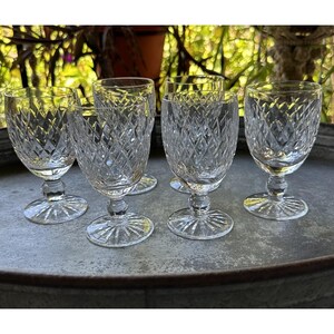 May include: Six clear crystal wine glasses with diamond-cut patterns. The glasses have a stemmed base and a round foot. The glasses are arranged on a metal tray, with a blurred background of green foliage.