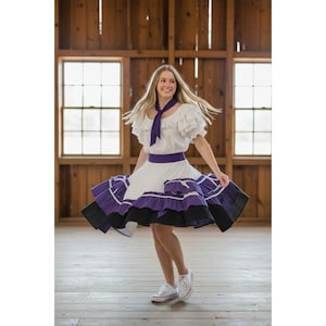 May include: A white blouse with ruffled sleeves and a purple neck scarf paired with a white skirt featuring a wide purple waistband and a multi-layered hemline with purple and black ruffles. The outfit is completed with white lace-up sneakers.